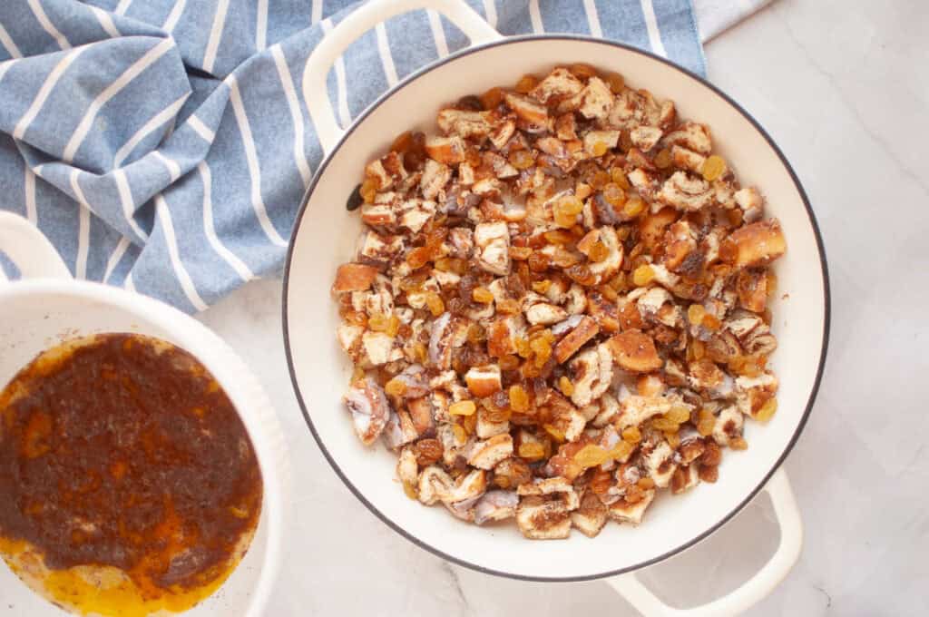 Overhead shot of chopped cinnamon rolls mixed with raisins in a white pot next to a bowl of wet ingredients.