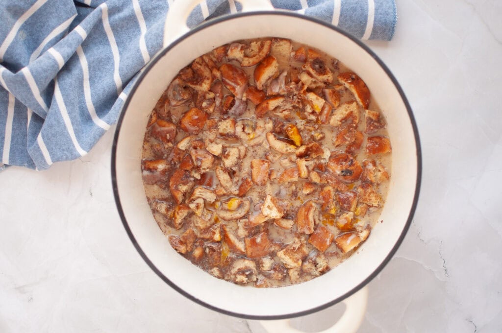 Overhead shot of chopped cinnamon rolls soaking in a light-colored custard mixture in a white pot.
