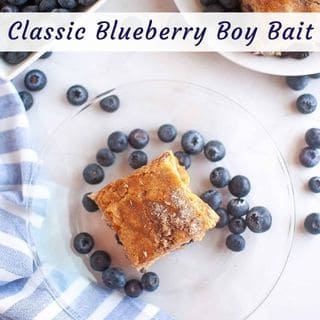 A square piece of Blueberry Boy Bait on a clear plate surrounded by blueberries, with a bowl of blueberries and more cake on a separate plate nearby.