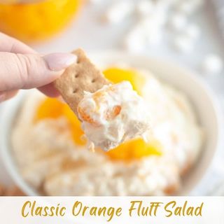 A hand holds a graham cracker dipped in creamy orange fluff salad with mandarin orange slices, with marshmallows visible in the background. Text reads "Classic Orange Fluff Salad.