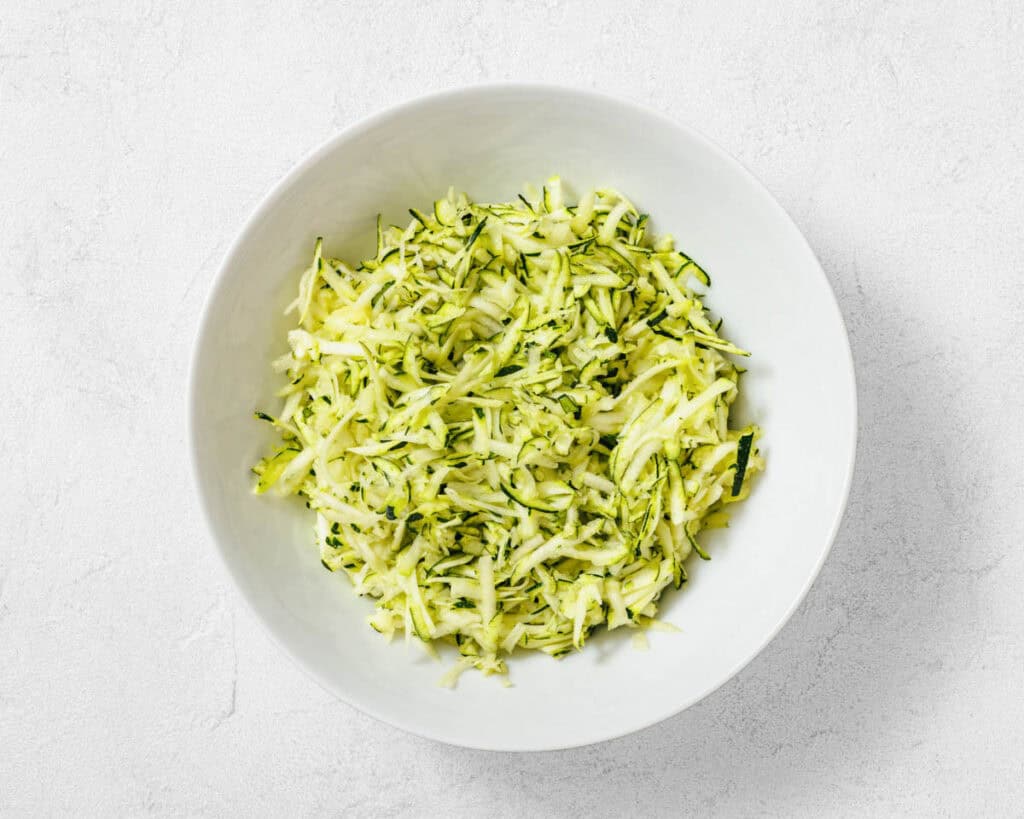 A white bowl filled with freshly grated zucchini on a light-colored surface.
