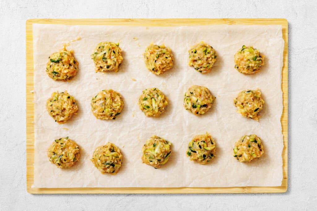 Raw vegetable and meat mixture formed into round portions arranged in rows on a parchment-lined wooden board, ready for baking or cooking.