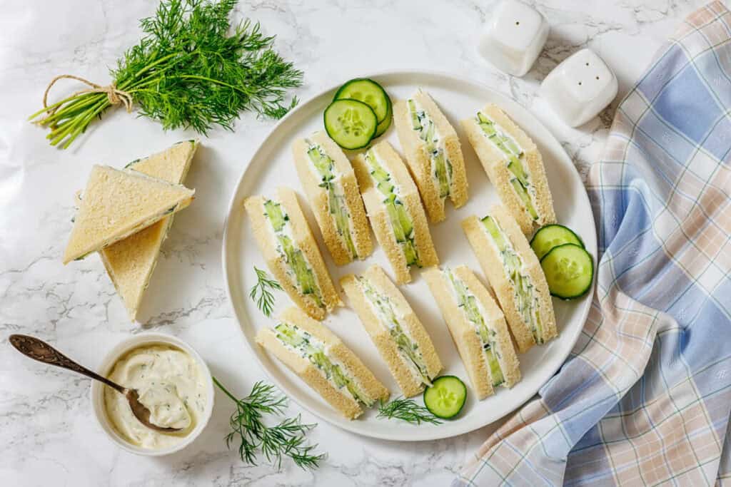 A plate of cucumber sandwiches with cream cheese, garnished with dill and cucumber slices, sits on a marble surface beside a bowl of spread, herbs, and a checked napkin.
