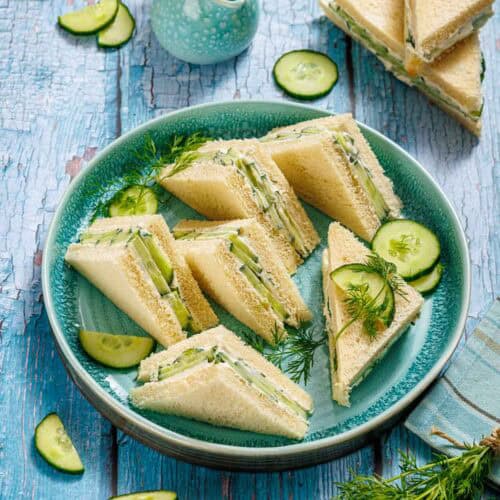 Triangle cucumber sandwiches with fresh dill and cucumbers are arranged on a blue plate, set on a rustic blue wooden table. Sliced cucumber and a small blue jug are nearby.