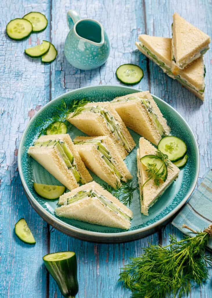 Triangle cucumber sandwiches with fresh dill and cucumbers are arranged on a blue plate, set on a rustic blue wooden table. Sliced cucumber and a small blue jug are nearby.