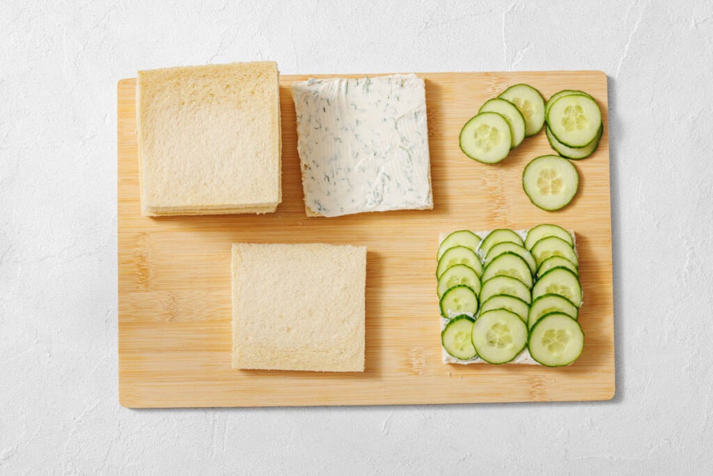 A wooden board with two slices of bread, one spread with herb cream cheese and cucumber slices, plus extra cucumber slices and bread stacked nearby.