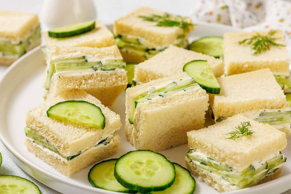 A plate of square cucumber tea sandwiches with cream cheese filling, garnished with cucumber slices and fresh dill.