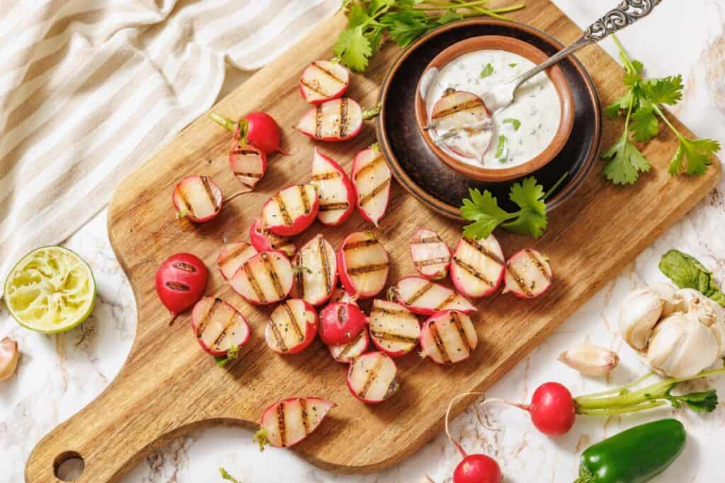 Grilled radish halves on a wooden board with a small bowl of creamy dipping sauce, garnished with cilantro, lime, garlic, and whole radishes.