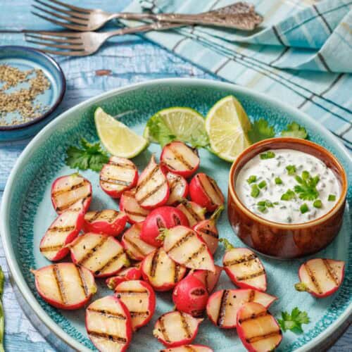 A plate of grilled radishes with lime wedges and a bowl of creamy dip, served on a blue ceramic dish with cutlery and a plaid napkin in the background.