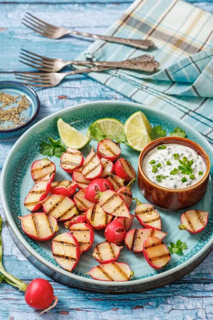A plate of grilled radishes with lime wedges and a bowl of creamy dip, served on a blue ceramic dish with cutlery and a plaid napkin in the background.