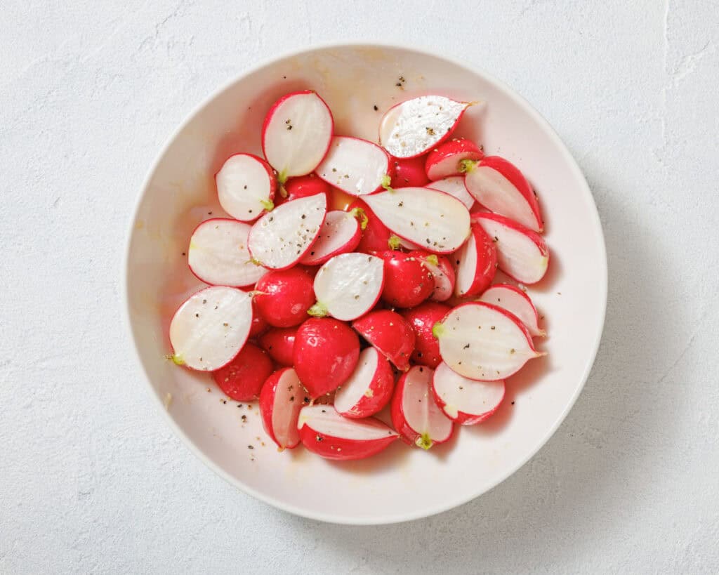 A white bowl filled with sliced radishes, lightly seasoned with salt and black pepper, on a light-colored surface.