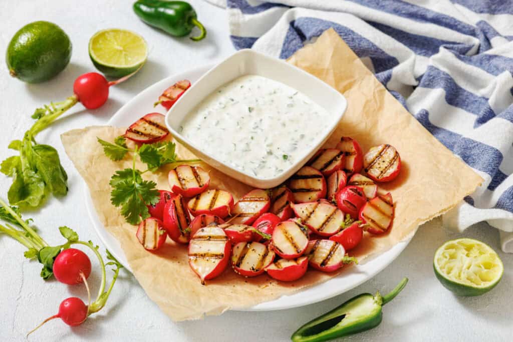 A plate of grilled radishes with a bowl of creamy dipping sauce, surrounded by fresh cilantro, lime halves, whole radishes, and jalapeños on a white surface.