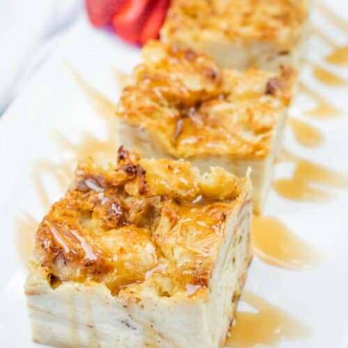 Three rectangular slices of bread pudding drizzled with caramel sauce on a white plate, garnished with a halved strawberry.