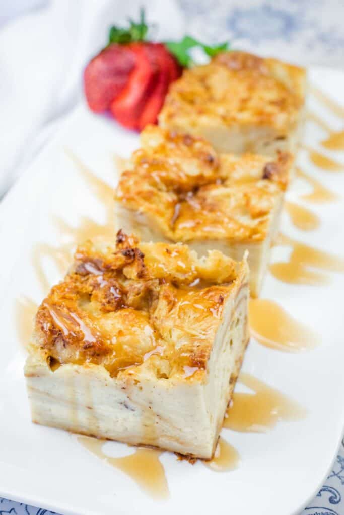 Three rectangular slices of bread pudding drizzled with caramel sauce on a white plate, garnished with a halved strawberry.