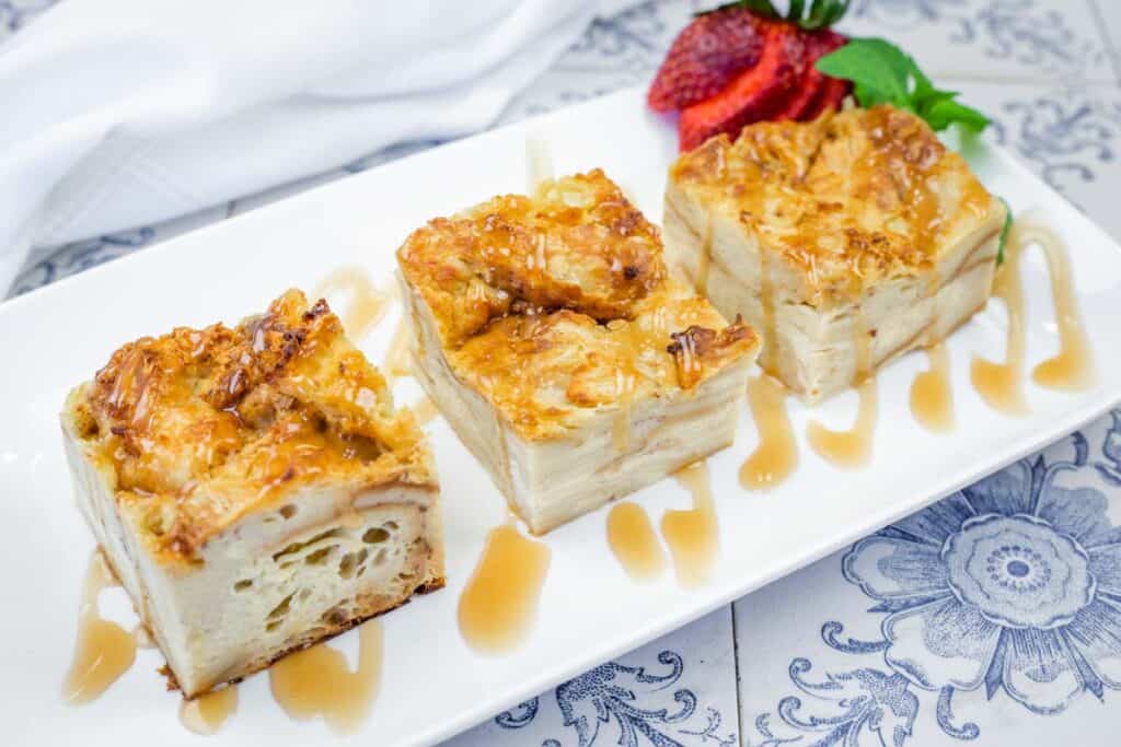 Three square pieces of bread pudding with caramel drizzle on a rectangular white plate, garnished with sliced strawberries and mint.