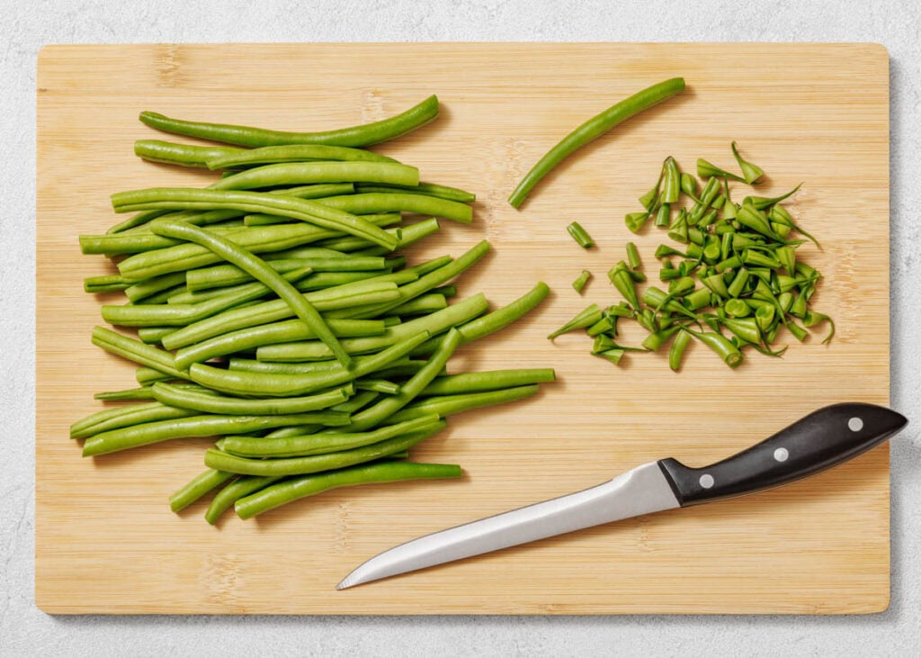 A pile of whole green beans, trimmed green bean tips, and a paring knife on a wooden cutting board.
