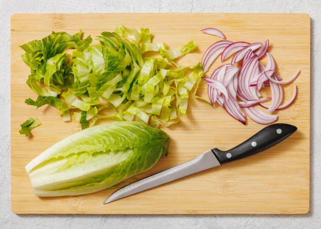 Chopped romaine lettuce, sliced red onion, and a knife on a wooden cutting board.