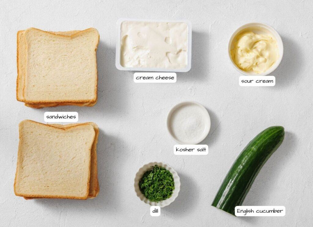 Four slices of sandwich bread, cream cheese, sour cream, kosher salt, chopped dill, and an English cucumber arranged on a white surface.