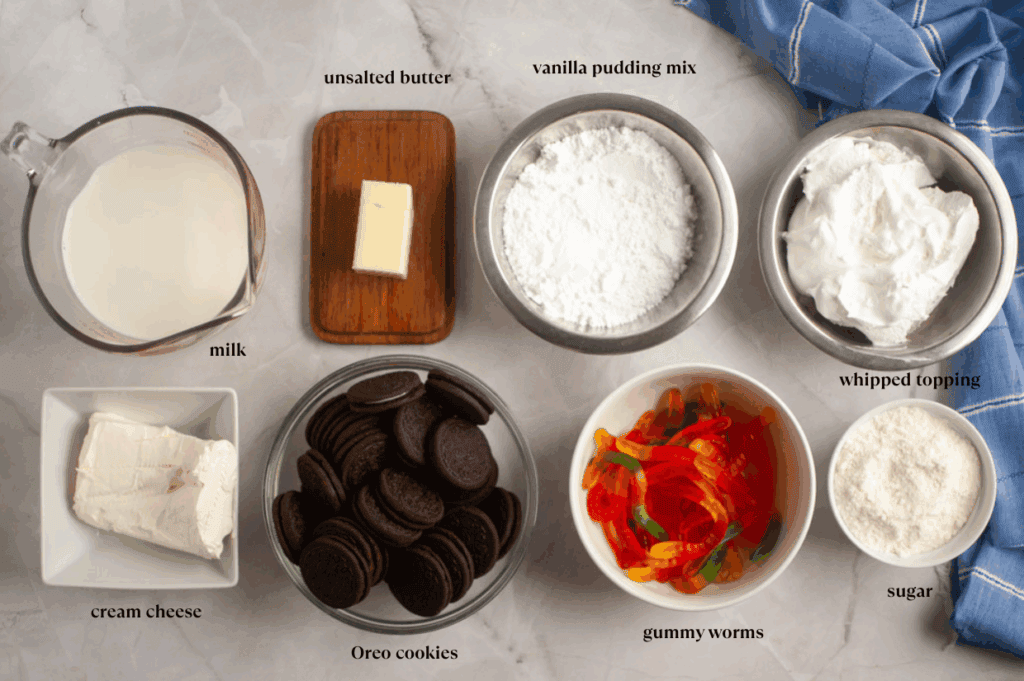 Ingredients for a dessert recipe arranged on a countertop: milk, unsalted butter, vanilla pudding mix, whipped topping, cream cheese, Oreo cookies, gummy worms, and sugar.