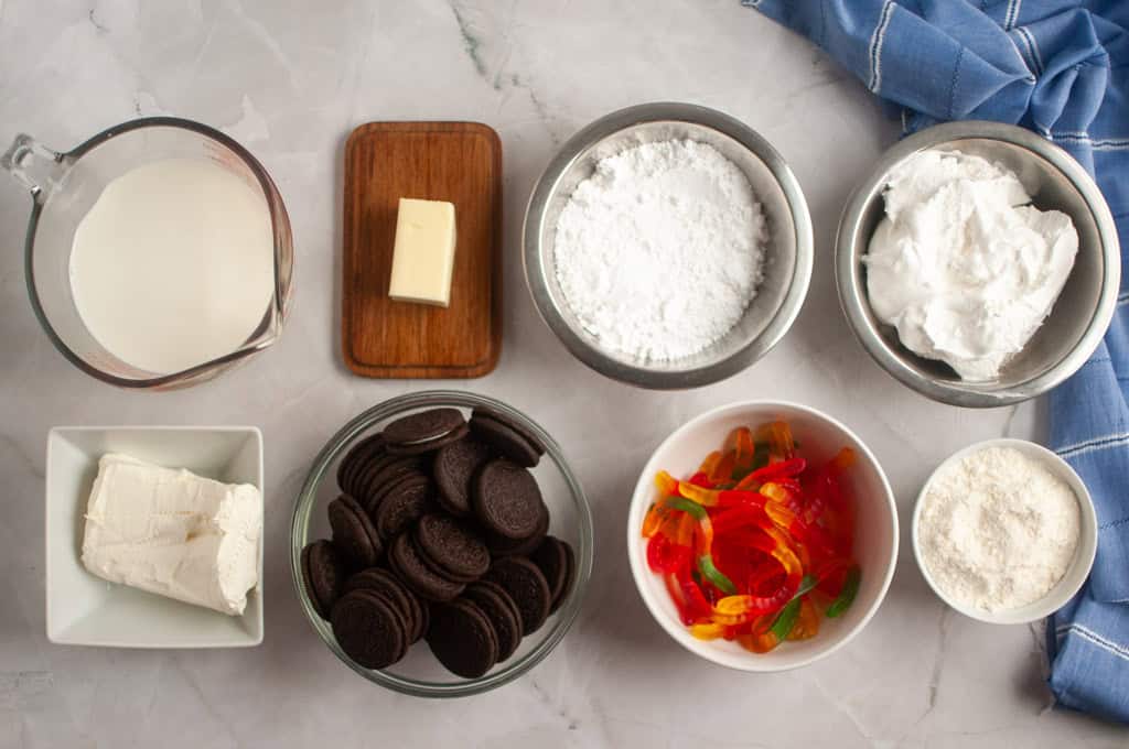 Assorted ingredients on a countertop, including milk, butter, powdered sugar, whipped topping, cream cheese, chocolate sandwich cookies, gummy worms, and shredded coconut.
