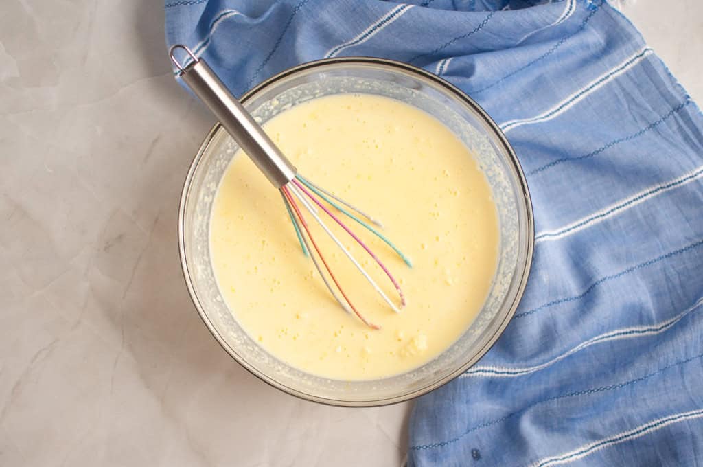 A glass bowl filled with a pale yellow batter and a whisk, placed next to a blue striped cloth on a light surface.
