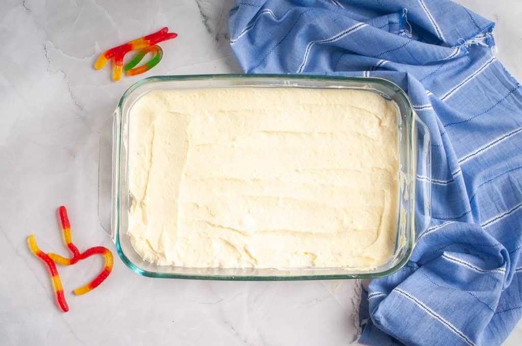 A glass baking dish filled with a smooth layer of pale batter next to a blue cloth and a few gummy worm candies on a marble surface.