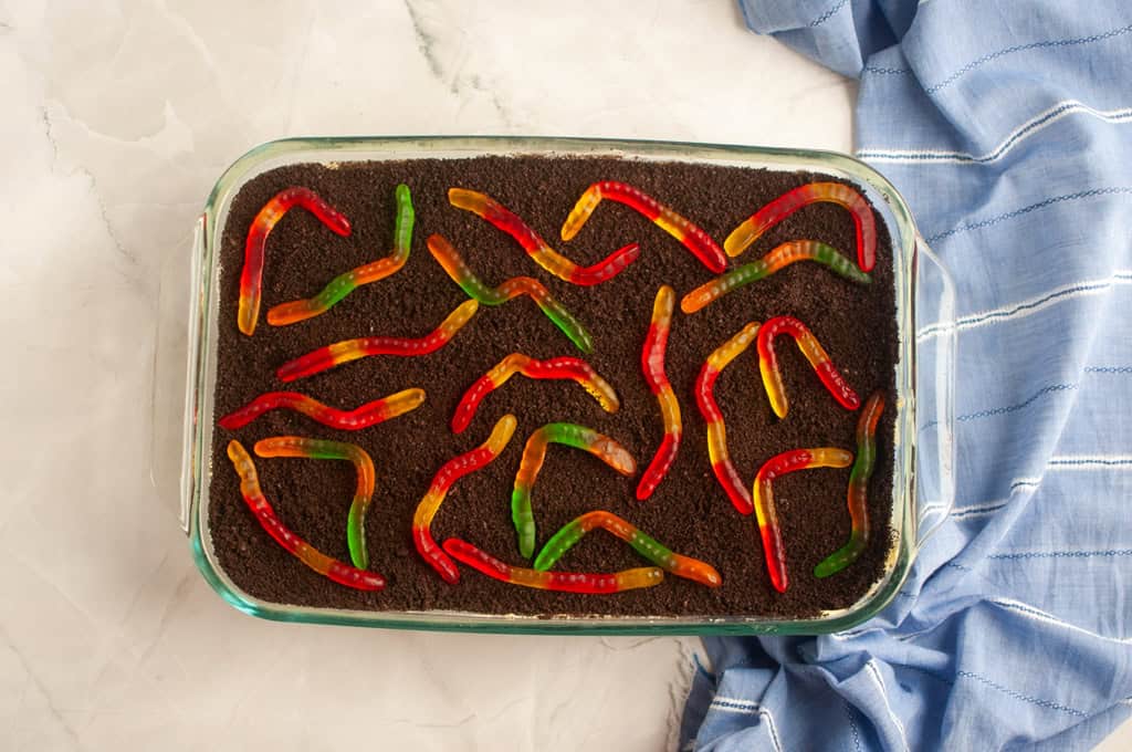 A glass baking dish filled with crumbled chocolate cookies topped with multi-colored gummy worms, next to a blue striped cloth on a marble surface.