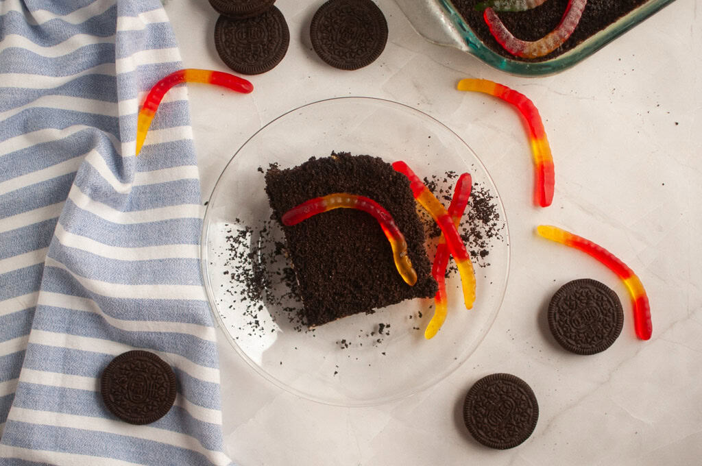 A slice of chocolate cake with crushed cookies and gummy worms on a clear plate, surrounded by whole cookies and a striped cloth.