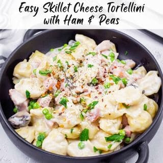 A black bowl filled with cheese tortellini, ham, and peas in a creamy sauce, garnished with herbs and black pepper.