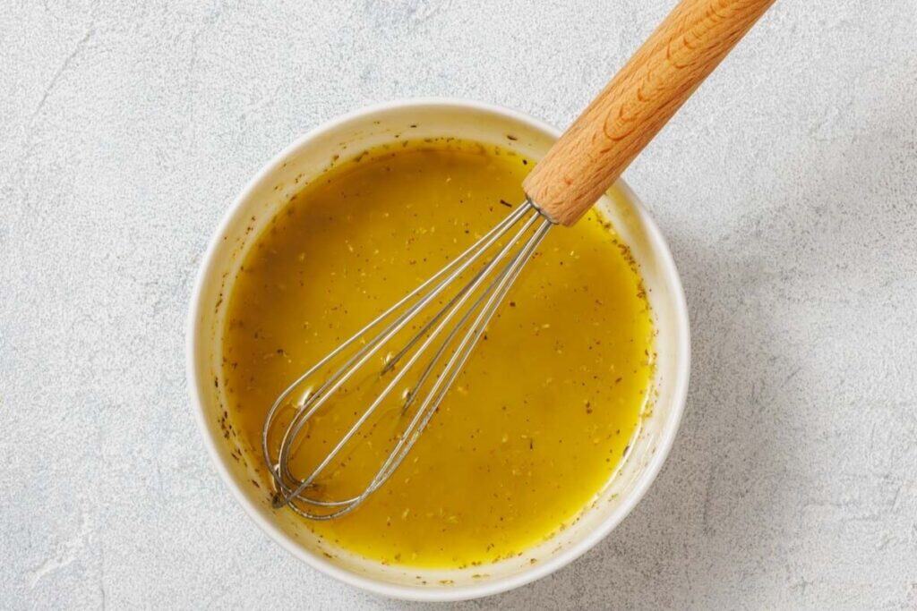 A white bowl filled with yellow salad dressing, being mixed with a metal whisk with a wooden handle, on a light surface.