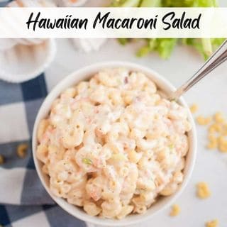 A white bowl filled with macaroni salad, containing elbow macaroni and a creamy dressing, is shown with a spoon inside. Celery and seashells are visible around the bowl.