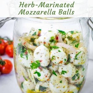 A glass jar filled with herb-marinated mozzarella balls, garnished with herbs and sliced onions. Some tomatoes are visible in the background.