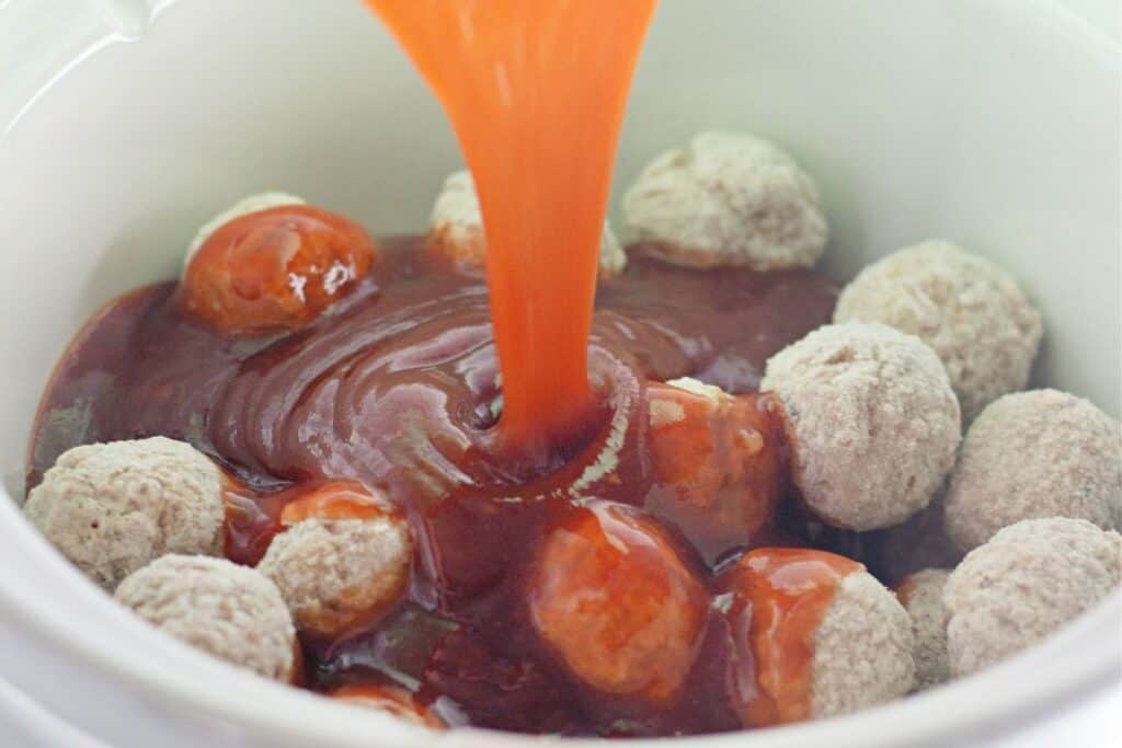 Frozen meatballs in a slow cooker with a red sauce being poured over them.