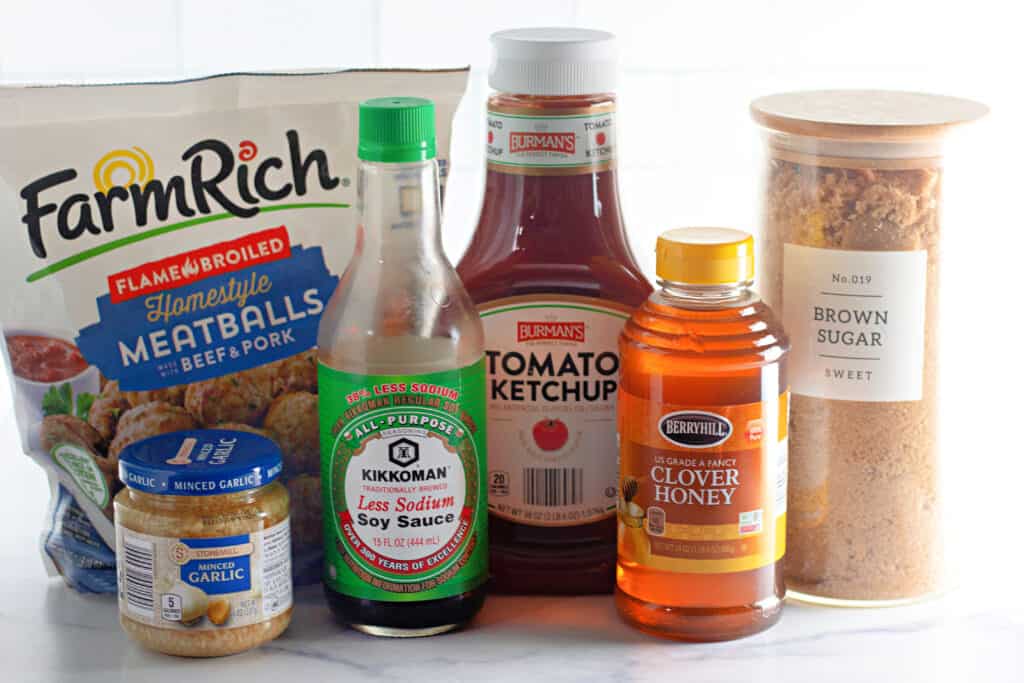 A collection of ingredients including packaged meatballs, minced garlic, soy sauce, ketchup, clover honey, and a container of brown sugar displayed on a white surface.