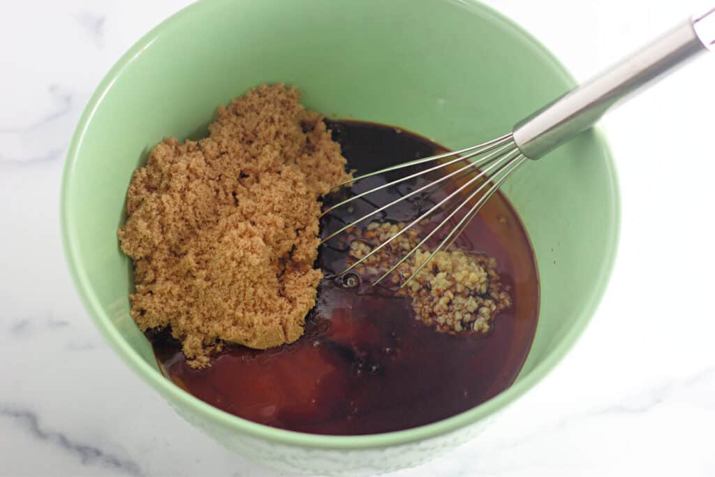 A green bowl containing brown sugar, minced garlic, and dark liquid ingredients with a metal whisk inside, set on a white surface.