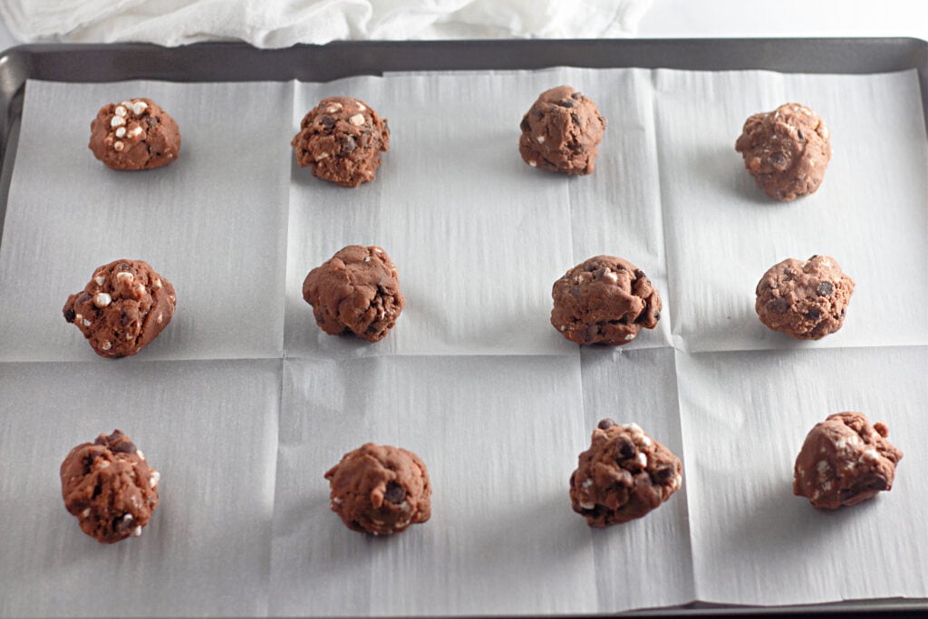 Twelve cocoa cookie dough balls arranged on a parchment-lined baking sheet, ready to be baked.