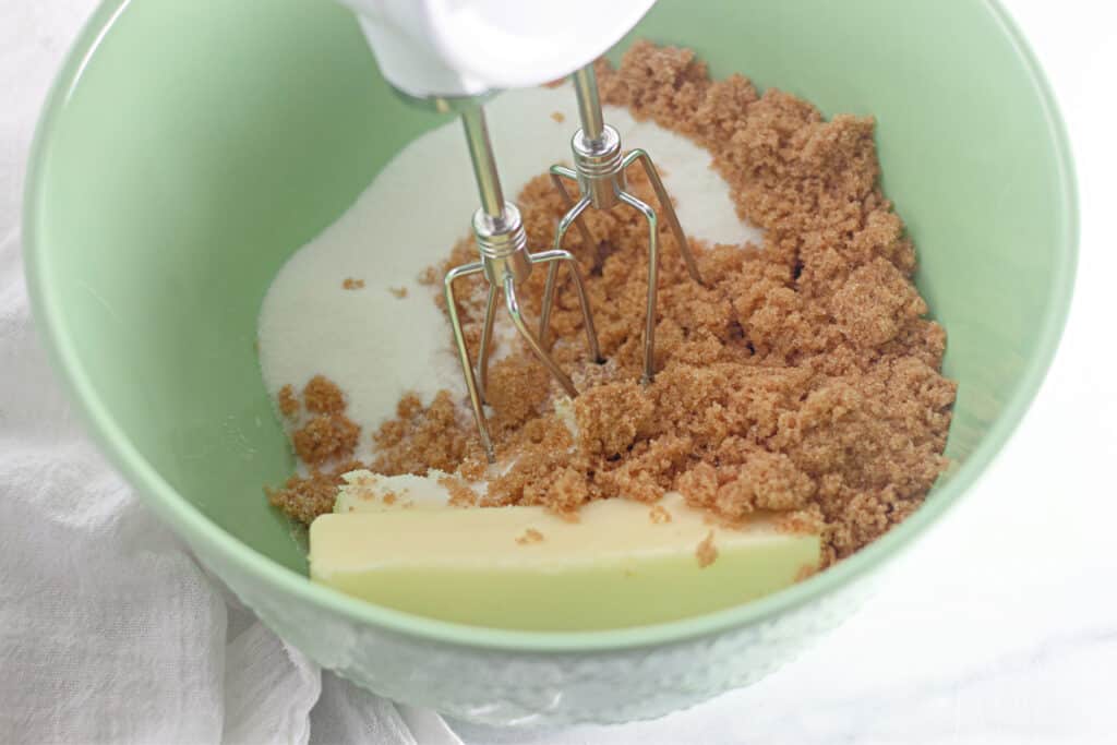 A mixing bowl with butter, granulated sugar, and brown sugar being mixed with an electric hand mixer.