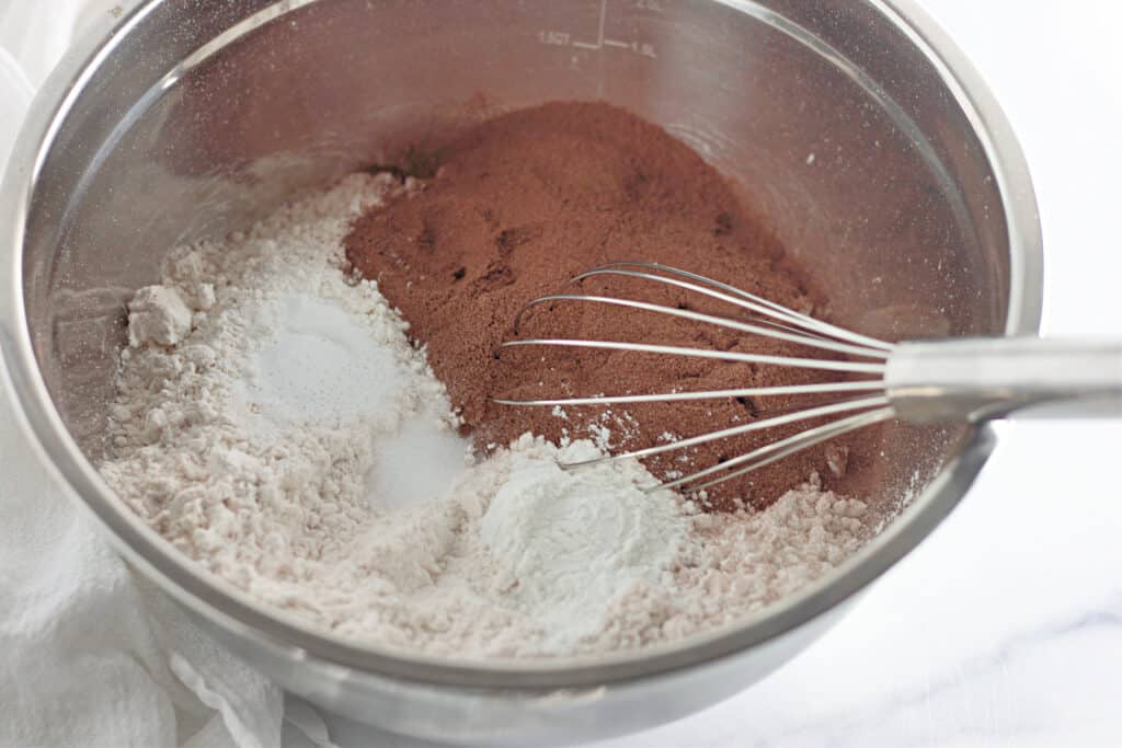 A metal mixing bowl with flour, cocoa powder, baking powder, and a metal whisk on a white surface.