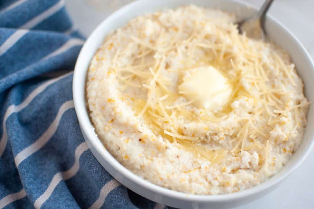 A white bowl filled with creamy grits, topped with shredded cheese and a melting pat of butter, with a spoon inserted.