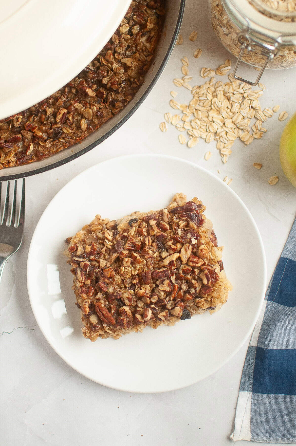 A slice of baked oatmeal topped with pecans on a white plate, with more in a baking dish.