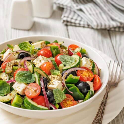 A bowl of Greek salad with tomatoes, cucumbers, red onions, olives, feta cheese, and mint leaves, placed on a white plate with a fork beside it. Salt and pepper shakers in the background.