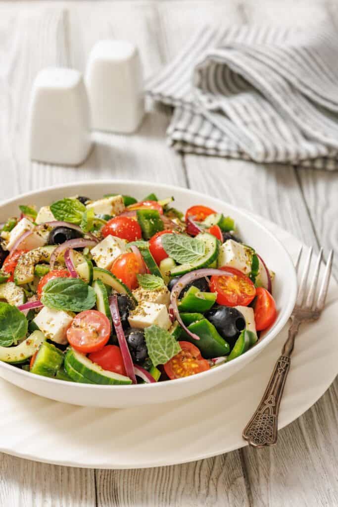 A bowl of Greek salad with tomatoes, cucumbers, red onions, olives, feta cheese, and mint leaves, placed on a white plate with a fork beside it. Salt and pepper shakers in the background.