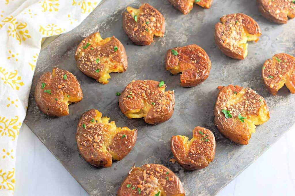 A baking sheet with several smashed roasted potatoes, garnished with herbs and grated cheese, arranged in rows.