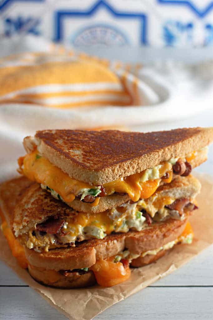 A stacked grilled cheese sandwich with melted cheddar, bacon, and creamy filling on toasted bread, placed on parchment paper with a cloth and bread loaf in the background.