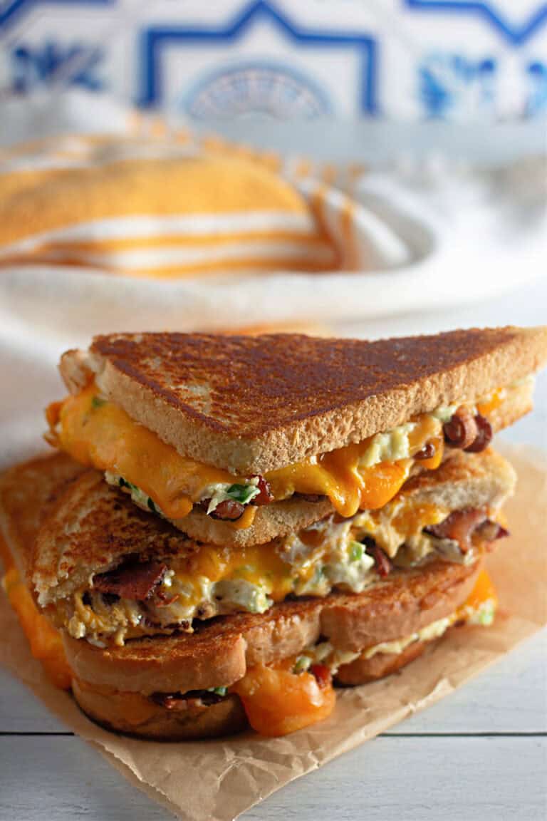 A stacked grilled cheese sandwich with melted cheddar, bacon, and creamy filling on toasted bread, placed on parchment paper with a cloth and bread loaf in the background.
