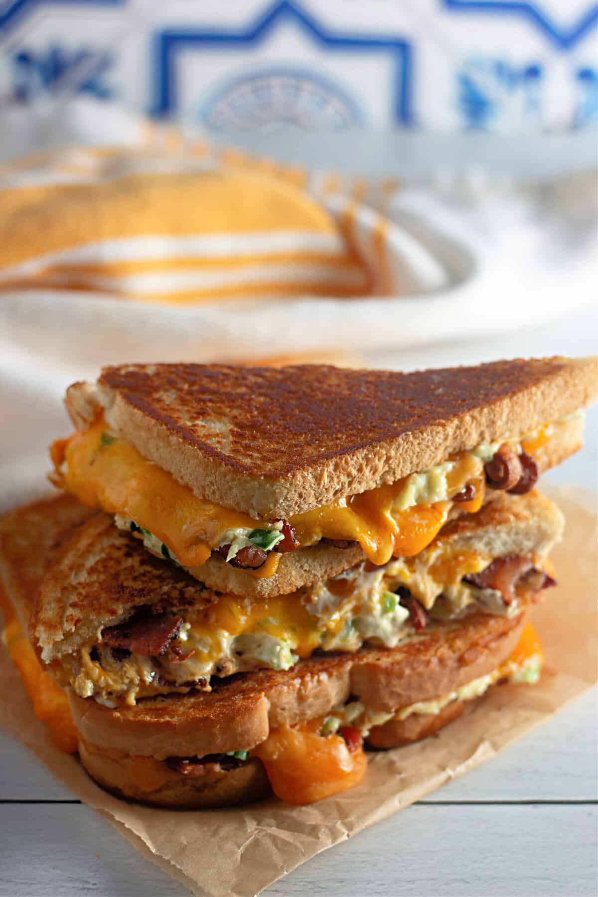 A stacked grilled cheese sandwich with melted cheddar, bacon, and creamy filling on toasted bread, placed on parchment paper with a cloth and bread loaf in the background.