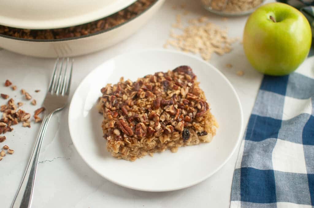 A slice of apple cinnamon baked oatmeal topped with pecans on a white plate with a fork.