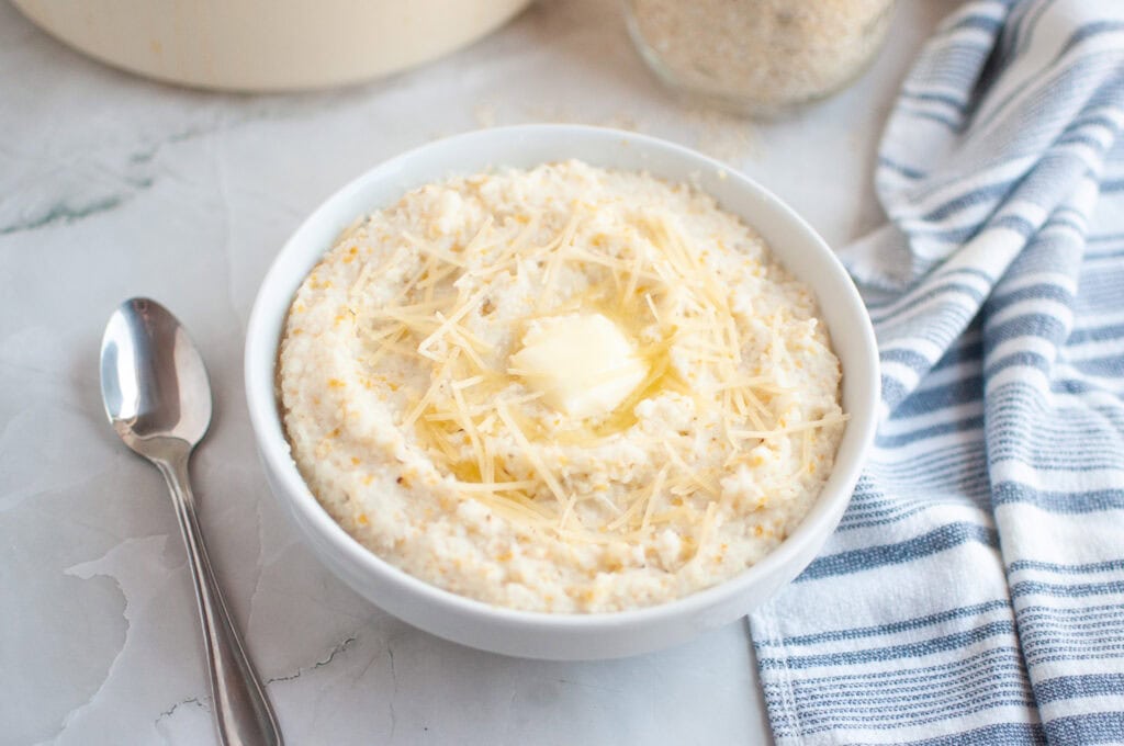 A white bowl of creamy grits topped with shredded cheese and a melting pat of butter, with a spoon beside it.