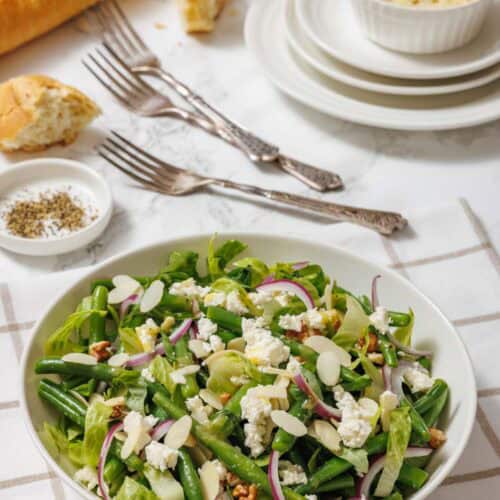 A bowl of green bean salad with feta, red onions, almonds, and pecans sits on a table with a baguette, white plates, forks, and a ramekin of slivered almonds.