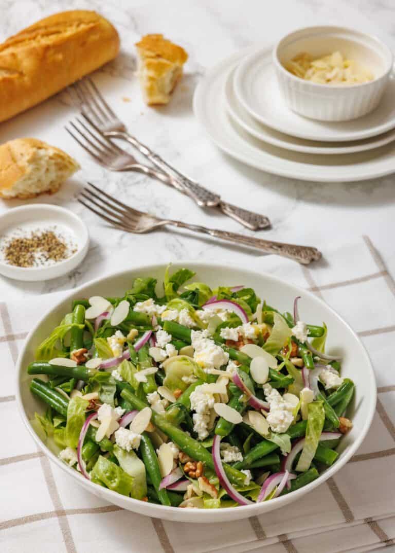 A bowl of green bean salad with feta, red onions, almonds, and pecans sits on a table with a baguette, white plates, forks, and a ramekin of slivered almonds.